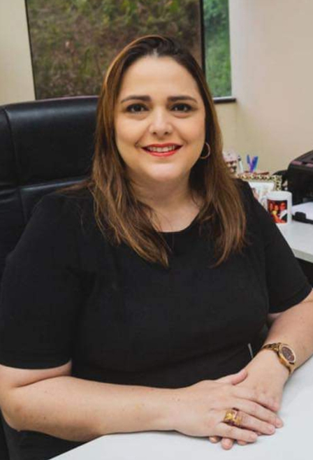 Na fotografia, mulher branca, com cabelos lisos, com mechas em tom castanho, olha e sorri para nós. Ela usa blusa preta, de mangas curtas, e está em ambiente com cadeira, mesa e equipamentos de escritório.