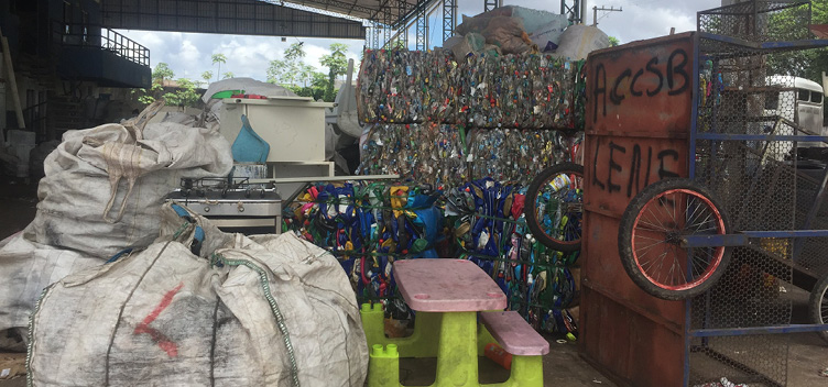 Fotografia contendo diversas sacas de lixo reciclável, à esquerda, uma estante com o lixo organizado, à direita. Em frente a estante, um carrinho de coleta de lixo e uma mesa de plástico com bancos em tamanho infantil.