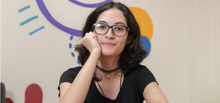 #ParaTodosVerem: Fotografia mostra uma mulher branca, jovem, de cabelos e olhos pretos. Ela usa óculos e veste uma blusa de manga curta, na cor preta. Ela está sentada, com o braço direito apoiado sobre um notebook; e o esquerdo, flexionado, com a mão apoiando o queixo. No fundo, uma parede de cor clara, ilustrada com desenhos coloridos e abstratos.