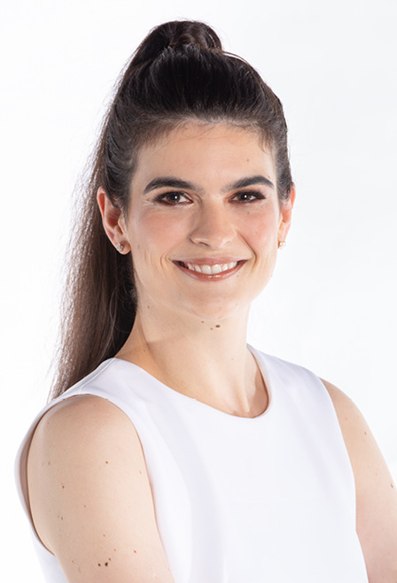  Fotografia mostra uma mulher branca. Ela tem cabelos e olhos castanhos, usa um vestido preto e branco, sem mangas, e está com os braços cruzados na altura da cintura. Os cabelos estão presos em forma de rabo de cavalo. Ela sorri em nossa direção.