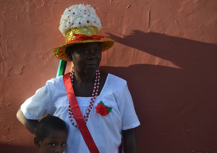 Fotografia de uma mulher negra. Ela olha em direção à câmera. Usa um traje típico da Marujada, composto por blusa branca, uma fita vermelha, colocada na diagonal do corpo. Do lado direito da blusa, há uma flor vermelha. No pescoço, colares de contas nas cores vermelha e branca. Ela usa também um chapéu de maruja.