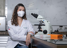 #ParaTodosVerem:Fotografia mostra uma mulher branca, jovem, de cabelos castanhos encaracolados, de comprimento médio. Ela usa máscara de proteção e veste um jaleco branco. Está sentada, apoiando os braços sobre a mesa. À sua direita, é possível ver um microscópio. Ao fundo, um quadro branco mostra anotações e gráficos.