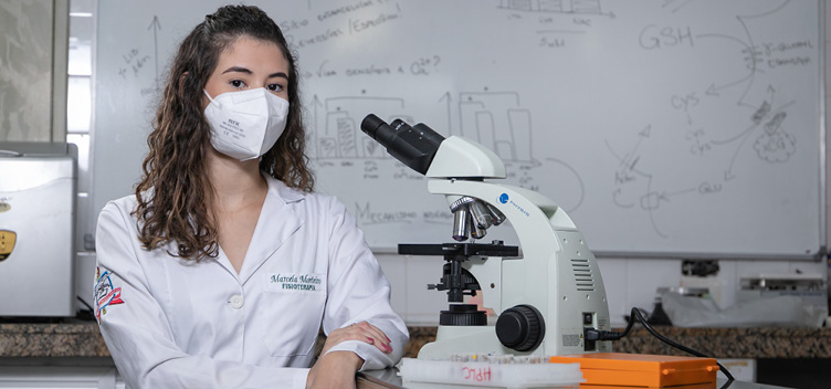 #ParaTodosVerem:Fotografia mostra uma mulher branca, jovem, de cabelos castanhos encaracolados, de comprimento médio. Ela usa máscara de proteção e veste um jaleco branco. Está sentada, apoiando os braços sobre a mesa. À sua direita, é possível ver um microscópio. Ao fundo, um quadro branco mostra anotações e gráficos.