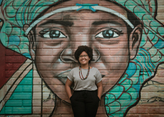 #ParaTodosVerem Fotografia mostra uma mulher negra, jovem, de cabelos castanhos e olhos pretos. Ela está sorrindo para nós. Veste calça na cor preta e blusa na cor bege. Usa óculos e colar colorido. Está com as costas apoiadas em uma parede ilustrada com grafite que retrata uma jovem negra.