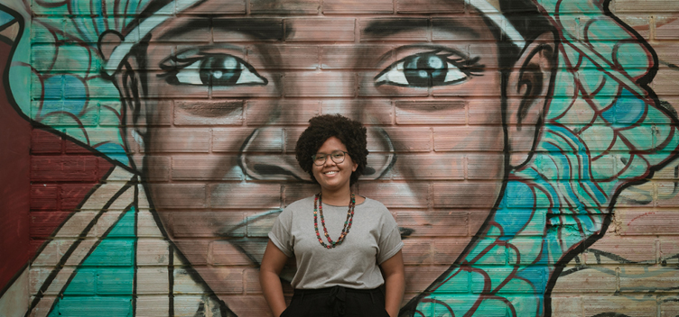 #ParaTodosVerem Fotografia mostra uma mulher negra, jovem, de cabelos castanhos e olhos pretos. Ela está sorrindo para nós. Veste calça na cor preta e blusa na cor bege. Usa óculos e colar colorido. Está com as costas apoiadas em uma parede ilustrada com grafite que retrata uma jovem negra.