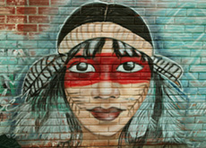#ParaTodosVerem: fotografia mostra grafite colorido que retrata o rosto de uma mulher indígena. Ela tem pintura corporal e usa um adorno com plumas na cabeça.