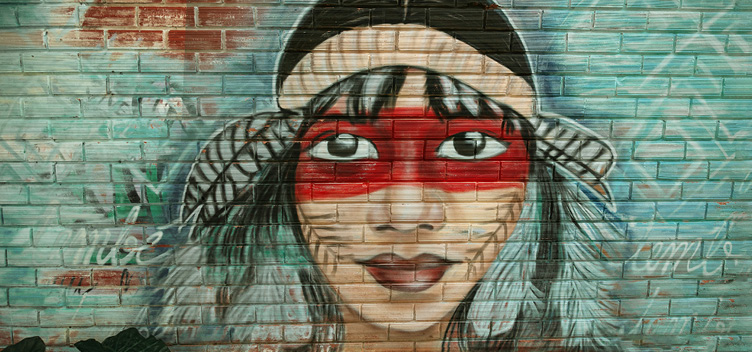 #ParaTodosVerem: fotografia mostra grafite colorido que retrata o rosto de uma mulher indígena. Ela tem pintura corporal e usa um adorno com plumas na cabeça.
