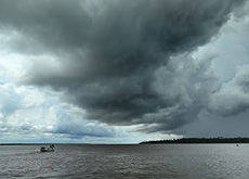 #ParaTodosVerem: Fotografia panorâmica em que se vê um pequeno barco navegando pelo rio. A vegetação ocupa as margens da direita e da esquerda, e o céu está coberto por nuvens de chuva.