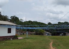 Fotografia da comunidade Nossa Senhora das Graças, Moju (PA), mostra um campo gramado, com construções em alvenaria nas laterais. O campo está ornamentado com bandeirolas nas cores branca e azul. No fundo, pequenas pontes dão acesso a casas de madeira. Por trás dessas casas, é possível ver a vegetação alta e fechada. 