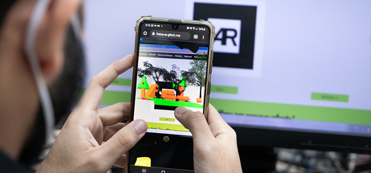 Fotografia mostra um homem usando um software de Realidade Aumentada no celular. O celular está centralizado na imagem e com a tela virada para nós. O monitor de um computador está ao fundo e com a tela virada para nós. No lado esquerdo da imagem, a cabeça do homem está desfocada. O homem está apontando a câmera do celular na direção de um código que está na tela do monitor. Na tela do celular, há, modelada em 3D, uma maquete de uma floresta.