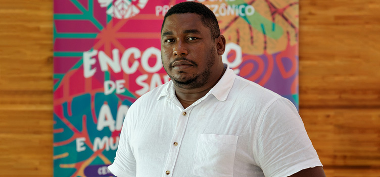 Davi Pereira Junior #ParaTodosVerem: Fotografia mostra um homem negro, de cabelos curtos, vestindo blusa branca. No fundo, um banner do 10º Fórum Social Pan-Amazônico.