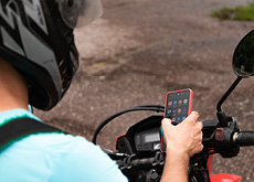 #ParaTodosLerem: Fotografia mostra um homem de costas para nós, sentado em uma motocicleta, olhando para a tela do seu celular. Ele usa um capacete preto, com detalhes na cor prata, e veste uma camiseta azul-clara, de manga curta.