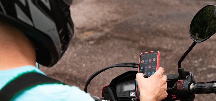 #ParaTodosLerem: Fotografia mostra um homem de costas para nós, sentado em uma motocicleta, olhando para a tela do seu celular. Ele usa um capacete preto, com detalhes na cor prata, e veste uma camiseta azul-clara, de manga curta.