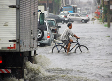 #ParaTodosLerem: Fotografia mostra pista inundada e trânsito desordenado. Em primeiro plano, veem-se três veículos enfileirados – um caminhão e dois automóveis de passeio - enfrentando o alagamento. Ao centro, tem-se a figura de um ciclista em posição perpendicular aos carros do primeiro plano. Ele veste blusa listrada e bermuda cinza. A água atinge as rodas da bicicleta até a altura da metade delas. Ao fundo, vê-se o fluxo de outros veículos enfrentando o alagamento e o horizonte completamente nublado. 