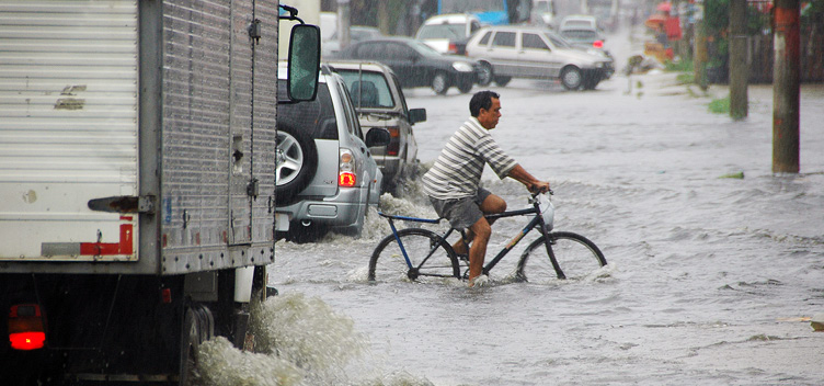 #ParaTodosLerem: Fotografia mostra pista inundada e trânsito desordenado. Em primeiro plano, veem-se três veículos enfileirados – um caminhão e dois automóveis de passeio - enfrentando o alagamento. Ao centro, tem-se a figura de um ciclista em posição perpendicular aos carros do primeiro plano. Ele veste blusa listrada e bermuda cinza. A água atinge as rodas da bicicleta até a altura da metade delas. Ao fundo, vê-se o fluxo de outros veículos enfrentando o alagamento e o horizonte completamente nublado. 