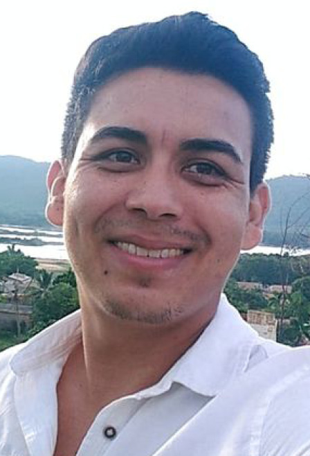 #ParaTodosLerem: Na fotografia de acervo pessoal, um homem pardo, com cabelos curtos, de cor preta, olha e sorri para nós. Ele usa camisa branca, de botão, e está em um ambiente ao ar livre, em que se destaca uma paisagem verde e um rio ao fundo.
