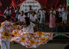 #ParaTodosLerem:  Fotografia mostra, em primeiro plano, um casal dançando carimbó. Ele veste calça branca e camisa estampada nas cores vermelha, laranja, amarela e branca. Ela veste blusa branca e saia rodada com a mesma estampa da camisa dele. Em segundo plano, um grupo canta e toca instrumentos de sopro e percussão. No fundo, uma bandeira vermelha em que se lê carimbó, patrimônio cultural.