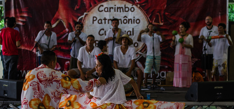 #ParaTodosLerem: Fotografia mostra, em primeiro plano, um casal dançando carimbó. Ele veste calça branca e camisa estampada nas cores vermelha, laranja, amarela e branca. Ela veste blusa branca e saia rodada com a mesma estampa da camisa dele. Em segundo plano, um grupo canta e toca instrumentos de sopro e percussão. No fundo, uma bandeira vermelha em que se lê carimbó, patrimônio cultural.