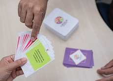 #ParaTodosLerem: Imagem apresenta, em ângulo fechado, um conjunto de cartas e três mãos em destaque. No canto esquerdo inferior, uma das mãos segura um grupo de cartas. Todas têm base de cor branca e centro coloridos. No canto esquerdo superior, a mão de uma segunda pessoa seleciona, utilizando o dedo indicador e o polegar, uma das cartas de centro vermelho. Em segundo plano, observam-se, apoiadas sobre uma mesa, duas pilhas de cartas. A pilha mais alta é composta por cartas de cor branca, com logotipo do produto ao meio. Uma pilha menor tem base de cor lilás, com losango branco ao centro, em que se observa a logo da coleção. Na ponta inferior direita da imagem, veem-se as pontas dos dedos da terceira mão sobre a mesa.