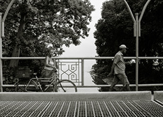 #ParaTodosLerem: Fotografia em preto e branco mostra duas pessoas atravessando a ponte sobre o rio Tucunduba, no campus da Universidade Federal do Pará, em Belém. À esquerda, uma mulher segura uma bicicleta pelo guidão enquanto anda. Ela veste short e camiseta, tem o cabelo preso e olha em nossa direção. À direita, o homem anda olhando para frente. Ele veste bermuda, camisa de manga curta e um boné. Ao fundo, veem-se a vegetação e o rio Guamá.