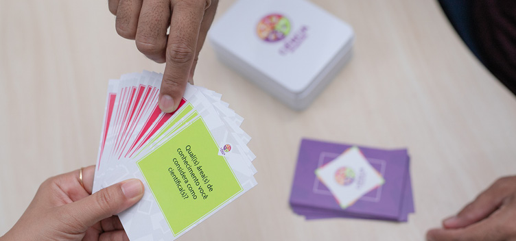 O conjunto de 45 cartas pode ser usado com alunos de diferentes níveis de formação, com o objetivo de suscitar debates sobre ciência e produção de conhecimento. #ParaTodosLerem: Imagem apresenta, em ângulo fechado, um conjunto de cartas e três mãos em destaque. No canto esquerdo inferior, uma das mãos segura um grupo de cartas. Todas têm base de cor branca e centro coloridos. No canto esquerdo superior, a mão de uma segunda pessoa seleciona, utilizando o dedo indicador e o polegar, uma das cartas de centro vermelho. Em segundo plano, observam-se, apoiadas sobre uma mesa, duas pilhas de cartas. A pilha mais alta é composta por cartas de cor branca, com logotipo do produto ao meio. Uma pilha menor tem base de cor lilás, com losango branco ao centro, em que se observa a logo da coleção. Na ponta inferior direita da imagem, veem-se as pontas dos dedos da terceira mão sobre a mesa.