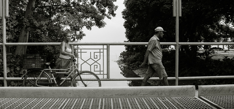 #ParaTodosLerem: Fotografia em preto e branco mostra duas pessoas atravessando a ponte sobre o rio Tucunduba, no campus da Universidade Federal do Pará, em Belém. À esquerda, uma mulher segura uma bicicleta pelo guidão enquanto anda. Ela veste short e camiseta, tem o cabelo preso e olha em nossa direção. À direita, o homem anda olhando para frente. Ele veste bermuda, camisa de manga curta e um boné. Ao fundo, veem-se a vegetação e o rio Guamá.