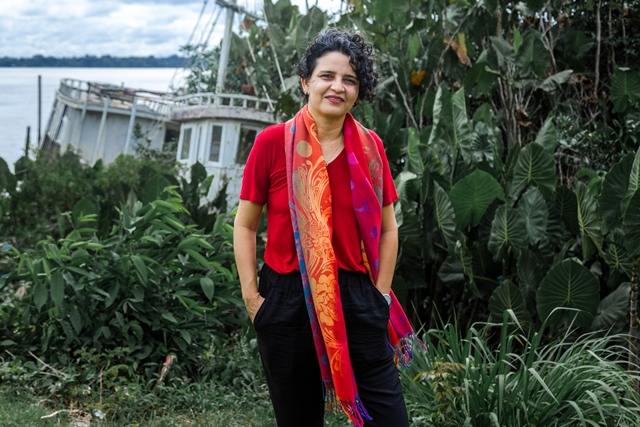 #ParaTodosVerem: Fotografia mostra, em primeiro plano, uma mulher de cabelos curtos e encaracolados, enquadrada do joelho para cima. Ela veste camiseta de mangas curtas, na cor vermelha, calça preta e carrega uma echarpe colorida no pescoço. Ela está de pé, com as mãos nos bolsos e sorri. Ao fundo, tem-se uma vegetação verde, uma embarcação ancorada e o rio Guamá.