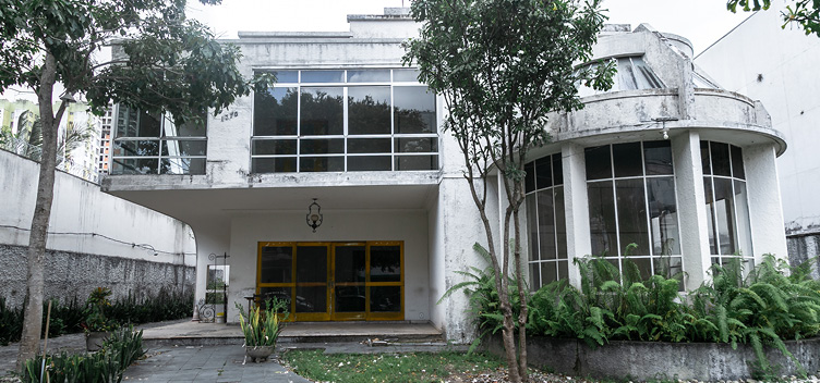 O Laboratório de Historiografia da Arquitetura e Cultura Arquitetônica (Lahca) da UFPA tem analisado a obra do engenheiro e arquiteto Camillo Porto de Oliveira. São construções, reformas e ampliações produzidas entre as décadas de 1940 e 1970. A Casa Moura Ribeiro faz parte desse acervo. #ParaTodosVerem: Fotografia da fachada de uma residência representante do estilo moderno de arquitetura. A estrutura tem dois pisos e apresenta pintura na cor branca, mas com marcas de ação do tempo. À esquerda, acima, destacam-se as vidraças que ocupam boa parte da fachada frontal e lateral da residência. Abaixo, há um pátio e uma porta também em vidraçaria, com detalhes na cor amarela. À direita, uma estrutura arqueada, apoiada sobre colunas e envolta por vidraças na frente e na lateral direita. À frente da residência, o calçamento é feito de pedras, com a presença de algumas plantas espalhadas e um jardim de samambaias, o qual envolve a base da estrutura arqueada da residência.