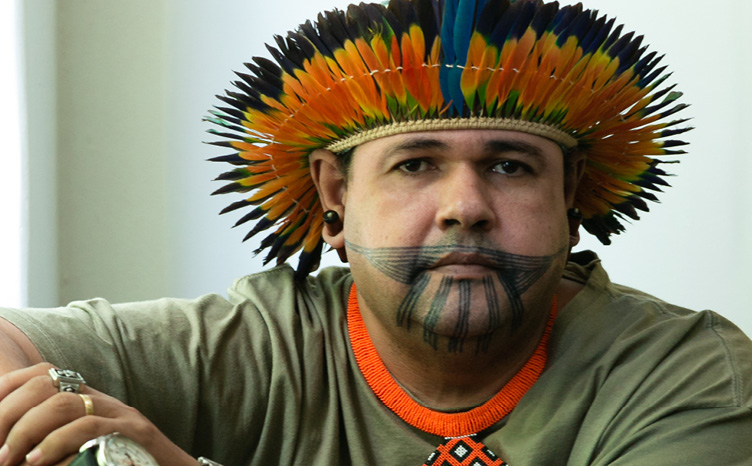 #ParaTodosVerem: Fotografia mostra um homem indígena sentado. Ele apoia o braço direito na janela e cruza o esquerdo a sua frente, segurando o próprio punho. Ele veste camisa de algodão na cor verde, usa relógio, anéis e pulseiras. Usa também um colar com grafismo indígena nas cores branca, preta e laranja. Sobre a sua cabeça, vemos um cocar. Em seu rosto, uma pintura corporal cobre os lábios, o queixo e parte da bochecha. Ele olha em nossa direção.