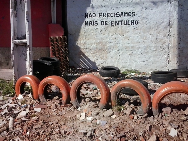 #ParaTodosVerem: Fotografia colorida mostra ponto de descarte irregular de resíduos sólidos no bairro Marambaia. Local traz pneus abandonados e sobras de construção civil. Pintura em muro ao fundo apresenta o texto: Não precisamos mais de entulho.