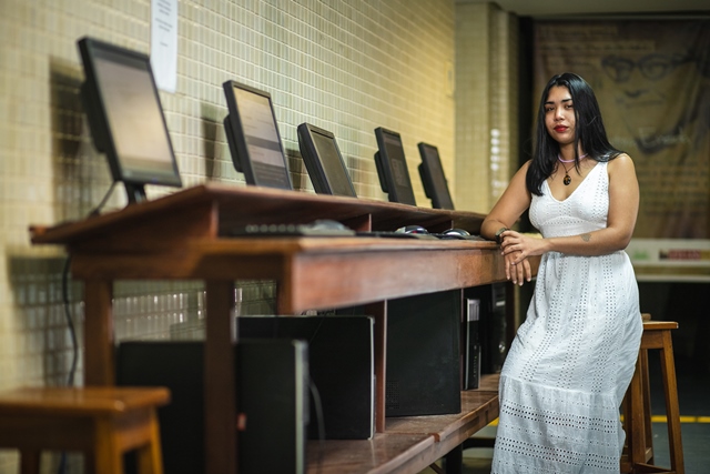 #ParaTodosVerem: Fotografia mostra uma mulher de cabelos lisos na altura dos ombros. Ela veste vestido longo, na cor branca, com decote em “v”. Ela está sentada em um banco alto, com um braço apoiado em uma bancada de madeira, onde vemos cinco computadores dispostos lado a lado. Ela olha em direção ao fotógrafo.