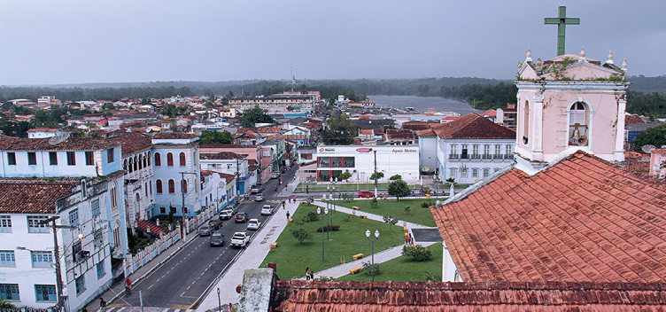 #ParaTodosVerem: Fotografia revela vista aérea do centro histórico da cidade de Bragança, no Pará. Em destaque, à direita e em primeiro plano, tem-se o telhado e a torre de uma igreja pintada em tom rosa. No alto da torre, há uma cruz na cor verde e um pequeno sino ligado a um megafone. À esquerda, destacam-se antigos casarões construídos em dois e três andares. À frente das edificações, observa-se uma avenida com movimento de veículos e pessoas e parte de uma praça. No horizonte, além da vista aérea de outras construções tem-se um rio e vegetação.