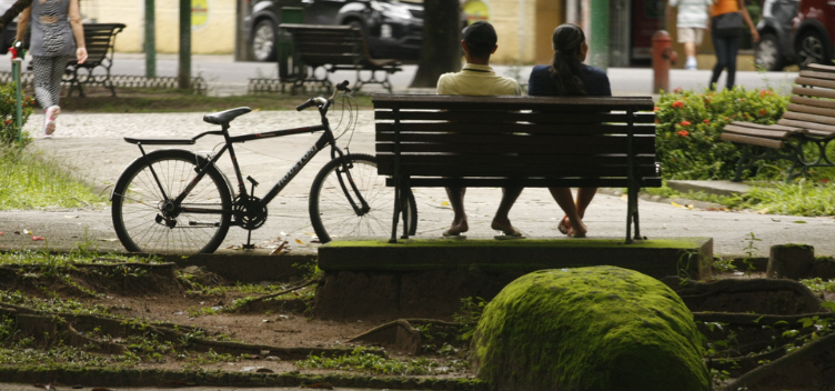 #ParaTodosVerem: Fotografia colorida mostra cena na praça Batista Campos. No centro da imagem, em destaque, aparece um casal de costas, sentado em um dos bancos da praça. Uma bicicleta encontra-se apoiada na ponta do mesmo banco. O homem veste camisa de cor amarelo-clara, usa boné e chinelo . A mulher tem cabelos longos, lisos e presos na altura da nuca. Ela usa blusa de cor azul-marinho e chinelos.