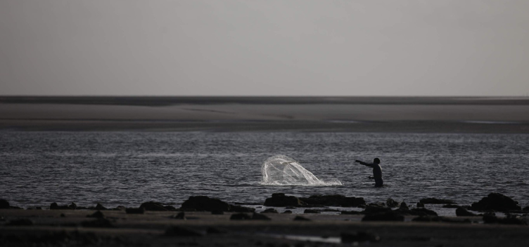 #ParaTodosVerem: Fotografia em preto e branco mostra paisagem litorânea. No rodapé da imagem, vemos uma faixa de areia e, em seguida, a formação de uma piscina natural. Na linha do horizonte, o banco de areia encontra com o céu. À esquerda da imagem, uma pessoa aparece dentro da água jogando uma rede de pesca.