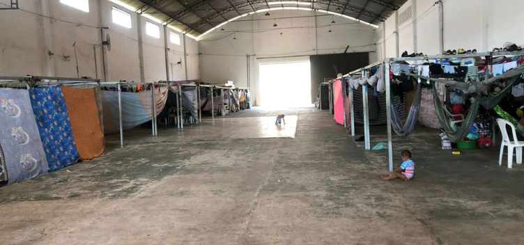 #ParaTodosVerem: Fotografia do espaço de acolhimento a imigrantes venezuelanos montado em Belém. Imagem destaca o espaço que se assemelha a um grande galpão, sem divisórias internas. Junto às paredes laterais, veem-se montadas diversas baias. A maioria delas está protegida por tecidos que fazem o papel de paredes individuais. Em outras, há redes atadas e roupas estendidas. Duas crianças brincam sobre o chão sem revestimento. No centro, ao fundo, há uma grande porta de correr que está aberta e deixa passar bastante claridade.