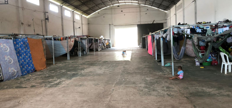 #ParaTodosVerem: Fotografia do espaço de acolhimento a imigrantes venezuelanos montado em Belém. Imagem destaca o espaço que se assemelha a um grande galpão, sem divisórias internas.  Junto às paredes laterais, veem-se montadas diversas baias. A maioria delas está protegida por tecidos que fazem o papel de paredes individuais. Em outras, há redes atadas e roupas estendidas. Duas crianças brincam sobre o chão sem revestimento.  No centro, ao fundo, há uma grande porta de correr que está aberta e deixa passar bastante claridade.