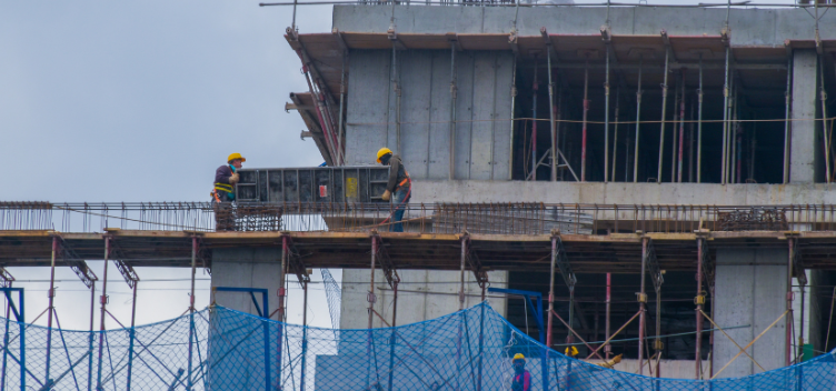 #ParaTodosVerem:A figura mostra um canteiro de obras com quatro trabalhadores. Eles usam capacetes amarelos, luvas, camisas de manga comprida e calças jeans. Dois deles estão sob uma estrutura de ferro e madeira e carregam uma placa de metal. Os outros dois, mais abaixo, conversam entre si. À frente deles, há uma rede de proteção na cor azul.