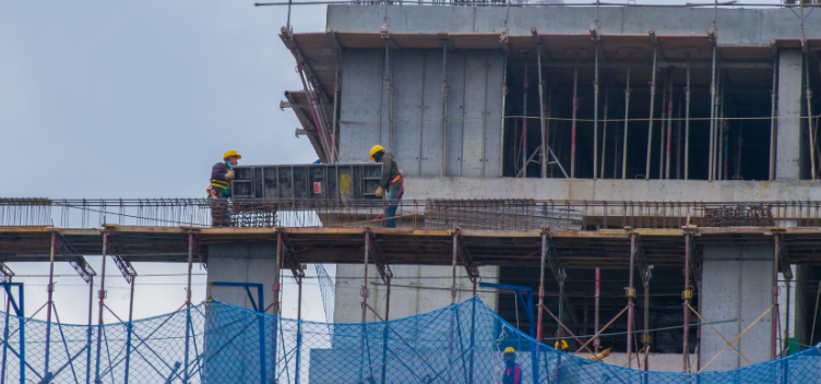 #ParaTodosVerem:A figura mostra um canteiro de obras com quatro trabalhadores. Eles usam capacetes amarelos, luvas, camisas de manga comprida e calças jeans. Dois deles estão sob uma estrutura de ferro e madeira e carregam uma placa de metal. Os outros dois, mais abaixo, conversam entre si. À frente deles, há uma rede de proteção na cor azul. 