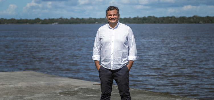 #ParaTodosVerem: Fotografia do professor da UFPA Pedro Aum. Ele está de pé, sobre uma base de concreto, à frente do rio Guamá. O professor usa camisa social na cor branca e calça cinza. Ele sorri em direção à câmera e suas mãos estão dentro do bolso da calça.