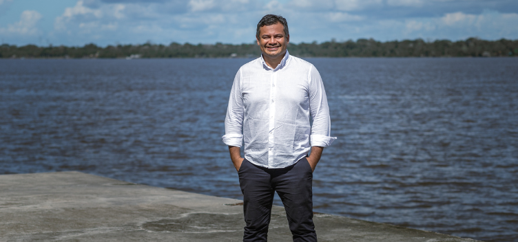 #ParaTodosVerem: Fotografia do professor da UFPA Pedro Aum. Ele está de pé, sobre uma base de concreto, à frente do rio Guamá. O professor usa camisa social na cor branca e calça cinza. Ele sorri em direção à câmera e suas mãos estão dentro do bolso da calça. 