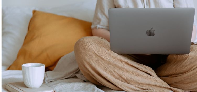 #ParaTodosVerem: Fotografia colorida mostra mulher sentada em uma cama com notebook no colo. Ela está enquadrada do ombro para baixo, veste calça larga bege e blusa creme. Ao seu lado há uma caneca de cerâmica branca apoiada sobre um pequeno livro de cor clara e uma travesseiro de fronha amarela. O lençol da cama é branco.
