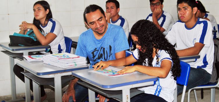 #ParaTodosVerem: Fotografia colorida mostra o ambiente de uma sala de aula. Em destaque, uma estudante faz uso de uma das maquetes táteis desenvolvidas durante a pesquisa para ensino adaptado de óptica. Ela esta sentada em uma carteira escolar e o material está sobre a mesa à sua frente. Ao seu lado, também sentado, está um professor auxiliar com outras maquetes táteis sobre a mesa. A sala tem outros estudantes.