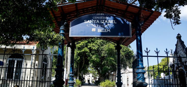 Localizado no bairro Guamá, o cemitério existe desde 1870 e possui mais de 80 mil túmulos. #ParaTodosVerem: Fotografia destaca fachada do Cemitério de Santa Izabel, na cor azul. Ao fundo, há árvores e várias lápides.