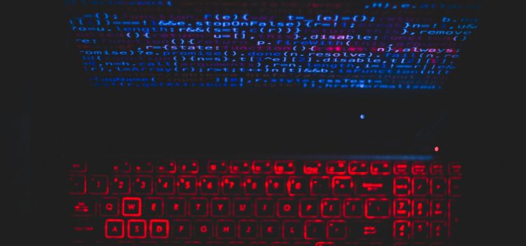 #ParaTodosVerem: A imagem apresenta fotografia de um computador notebook. No canto inferior, há um teclado iluminado por luz de neon vermelha. No canto superior, há uma tela com códigos de programação em tons de azul e rosa.