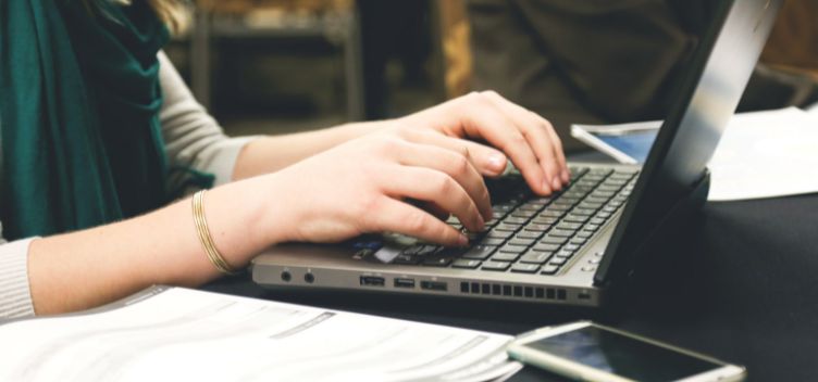 #ParaTodosVerem: A imagem mostra uma pessoa utilizando um notebook. Ao redor dele, aparecem um celular e folhas de papel.