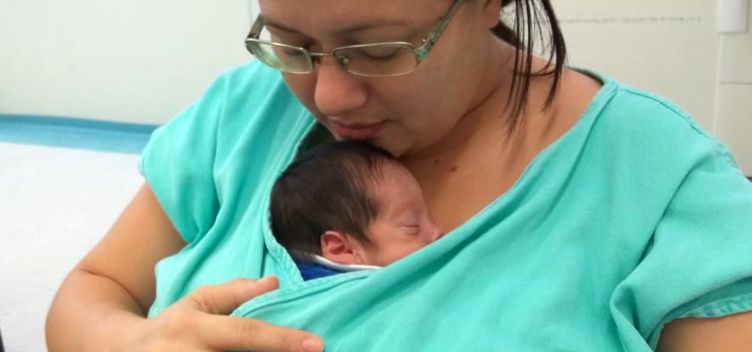 #ParaTodosVerem: Fotografia destaca uma mulher branca, de cabelos pretos, que carrega um recém--nascido sob a bata fornecida pelo hospital Santa Casa. Ela usa óculos, segura o bebê com a mão direita e apoia o queixo sobre a cabeça dele. A criança veste roupa de cor azul e parece dormir.