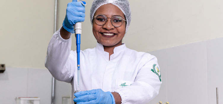 ParaTodosVerem: Fotografia colorida mostra jovem sorrindo. Ela usa equipamentos de segurança como touca e luvas descartáveis e jaleco branco. A jovem usa óculos de grau e nas mãos carrega instrumentos de laboratório utilizados para medição de líquidos.
