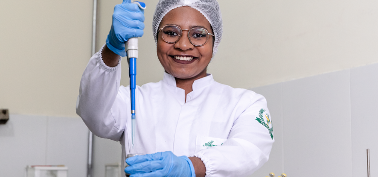 ParaTodosVerem: Fotografia colorida mostra jovem sorrindo. Ela usa equipamentos de segurança como touca e luvas descartáveis e jaleco branco. A jovem usa óculos de grau e nas mãos carrega instrumentos de laboratório utilizados para medição de líquidos. 