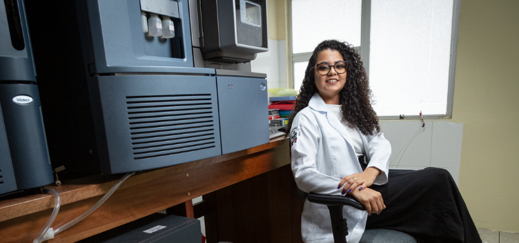 ParaTodosVerem: Fotografia colorida mostra jovem sorrindo ao lado de alguns maquinários de laboratório. Ela usa óculos, tem cabelo comprido, cacheado e de cor castanho-escuro. A jovem está sentada, tem as pernas e os braços cruzados e usa jaleco branco e calça preta.