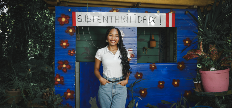 #ParaTodosVerem: Fotografia mostra mulher jovem, negra, de cabelos pretos, trançados. Ela veste blusa branca e calça jeans, está de pé e sorri para a câmera. Ao fundo, uma casa de madeira pintada de azul, com flores vermelhas. No alto da parede, há uma placa em que se lê: “sustentabilidade”. Ao redor da casa, há ampla vegetação.